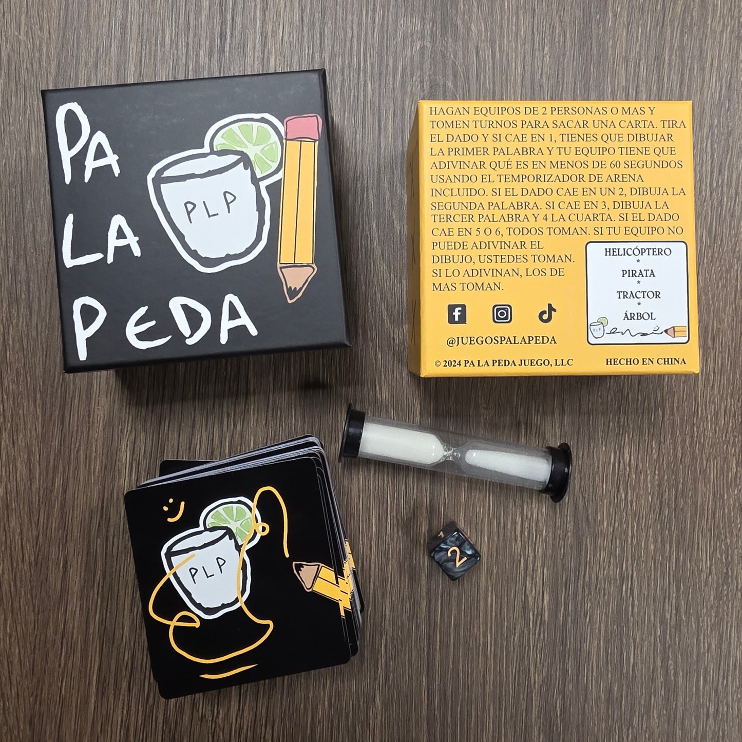 Two black boxes with white text and graphics on a wooden surface, with a yellow box and hourglass, cards and dice in the background.