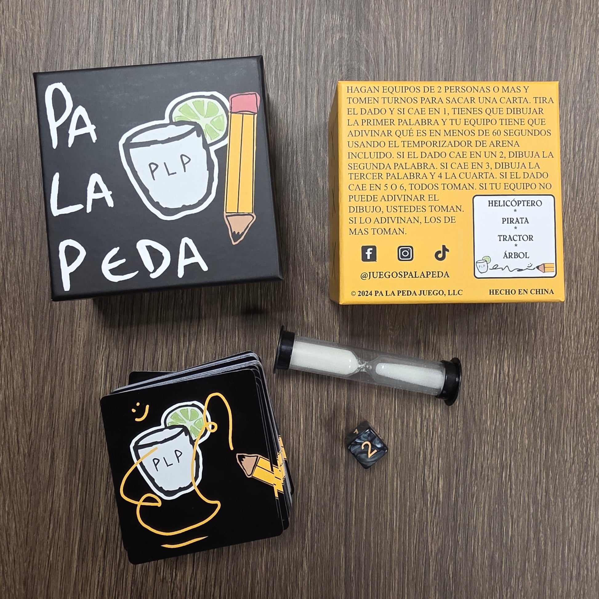 Two black boxes with white text and graphics on a wooden surface, with a yellow box and hourglass, cards and dice in the background.